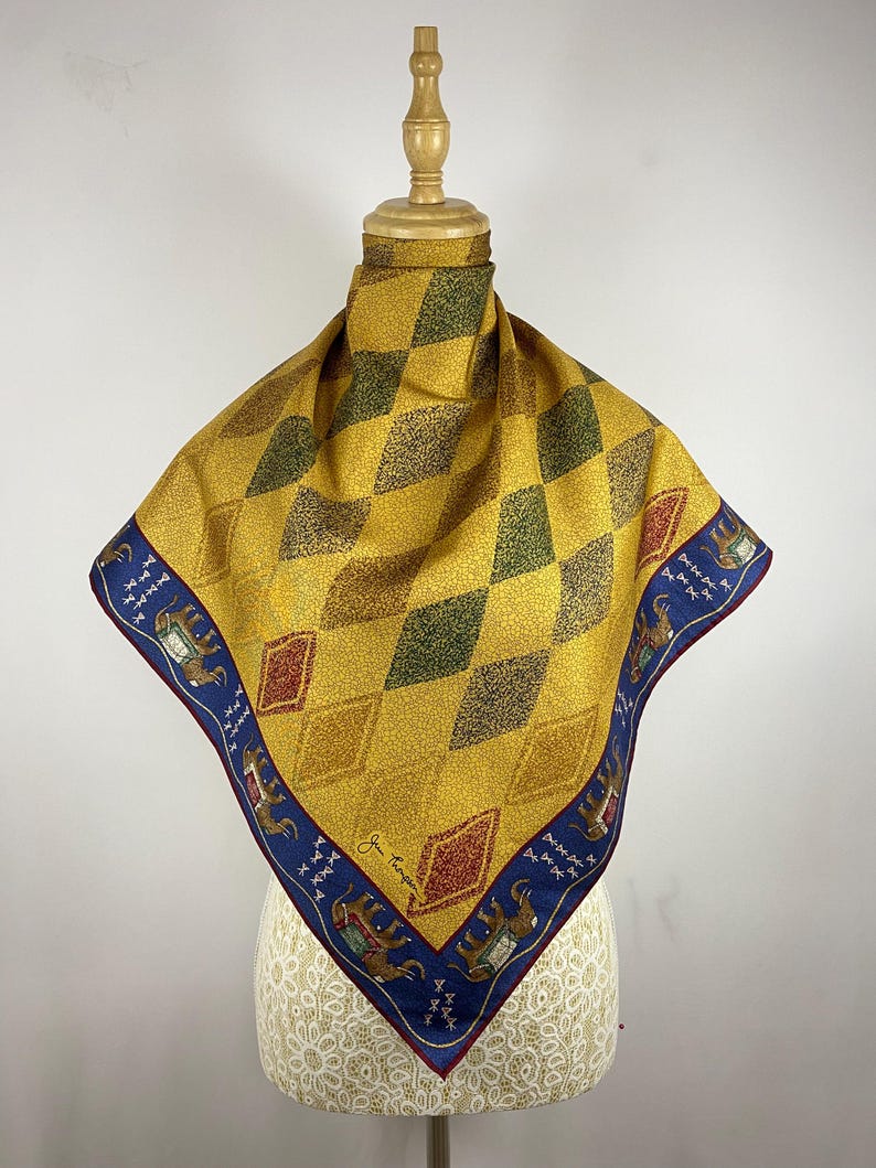 May include: A square scarf featuring a yellow and green diamond pattern across the center. The border is a deep blue color decorated with illustrations of elephants in ornate saddles and small white symbols. The signature Jim Thompson is visible.