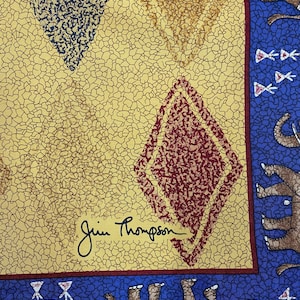May include: A fabric textile featuring a yellow background with a black crackle pattern and diamond shapes in red and dark blue. A blue border displays brown elephants and small white floral motifs. The signature Jim Thompson is printed in black.