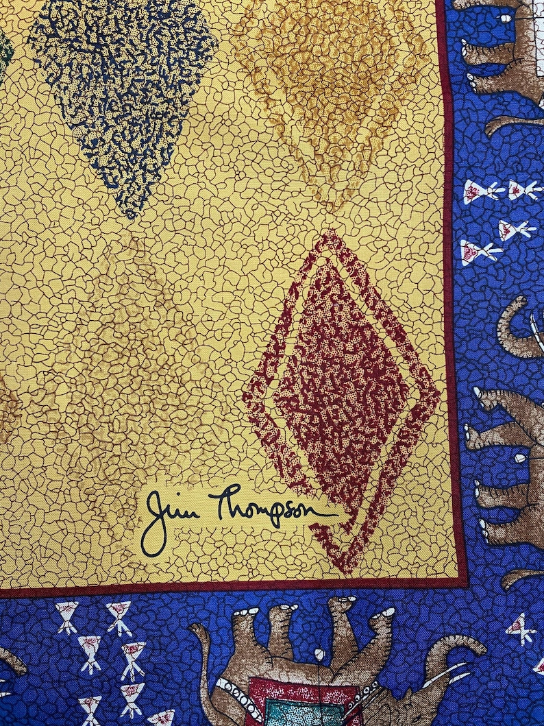 May include: A fabric textile featuring a yellow background with a black crackle pattern and diamond shapes in red and dark blue. A blue border displays brown elephants and small white floral motifs. The signature Jim Thompson is printed in black.