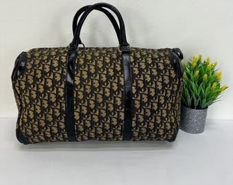May include: A brown and black patterned duffel bag with black leather handles and trim. The bag has a repeating pattern and a rectangular shape. The handles are arched and the bag appears to be made of a durable material.