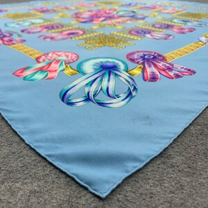 May include: A light blue silk scarf with a repeating pattern of colourful ribbons and bows. The scarf has a gold trim around the edges.