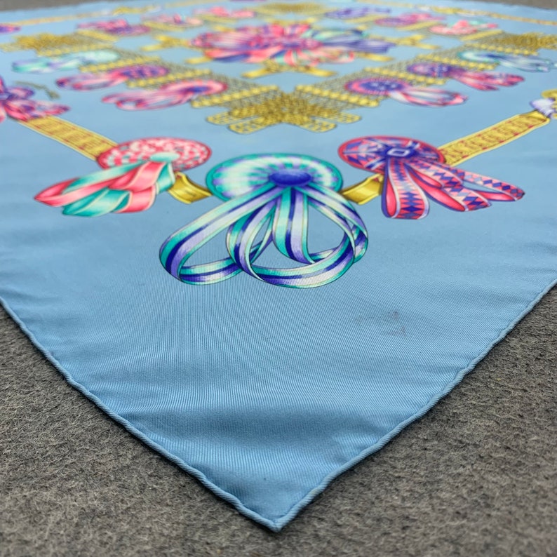 May include: A light blue silk scarf with a repeating pattern of colourful ribbons and bows. The scarf has a gold trim around the edges.