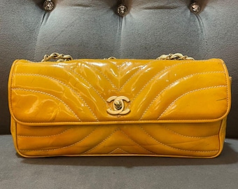 May include: A mustard-yellow, quilted leather handbag with a gold chain strap. The bag features a prominent interlocking logo on the front flap. The bag is rectangular and has a glossy finish, with a chevron pattern.