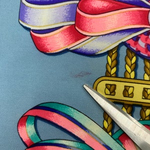 May include: A blue silk scarf featuring a colourful ribbon design. The ribbons are pink, blue, green and white. The scarf has a gold chain with a gold clasp.