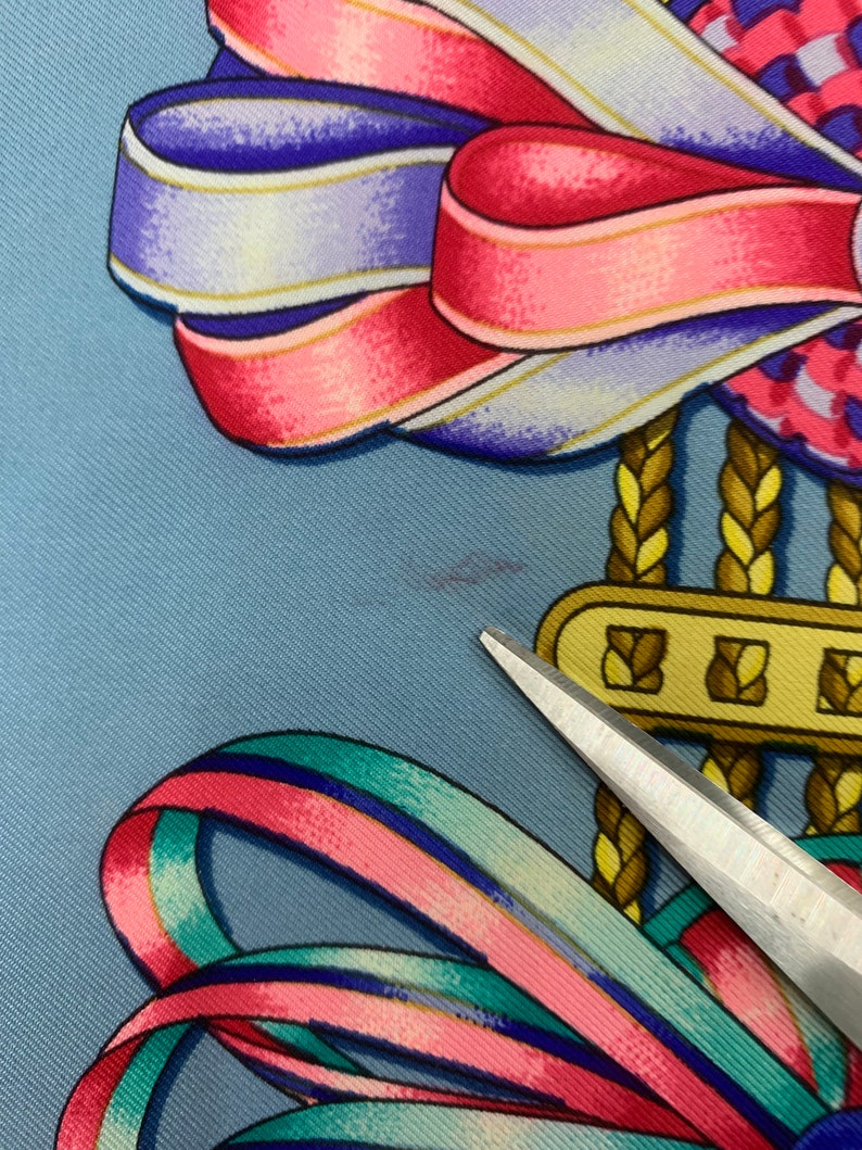 May include: A blue silk scarf featuring a colourful ribbon design. The ribbons are pink, blue, green and white. The scarf has a gold chain with a gold clasp.