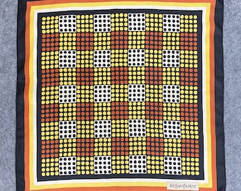May include: Square silk scarf with a geometric pattern of dots in yellow, orange, white, and black. The scarf has a black border with orange and yellow stripes. The brand name "YVES SAINT LAURENT" is printed in white.