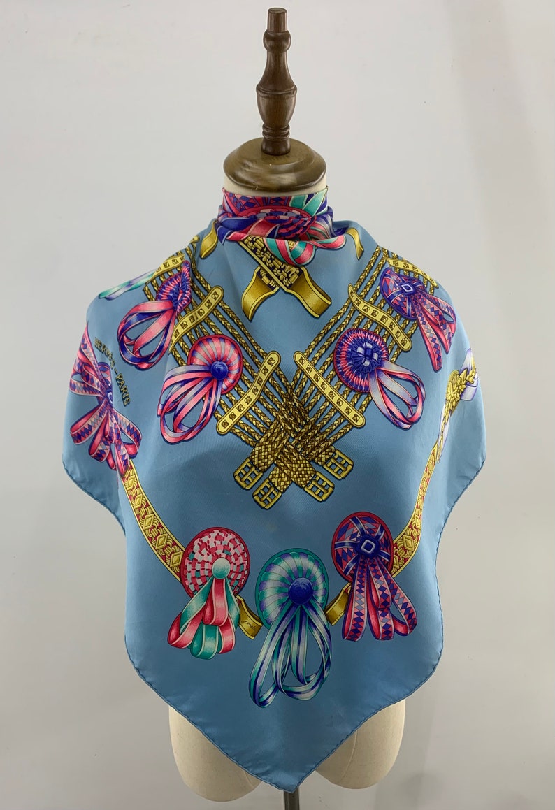 May include: A light blue silk scarf with a pattern of gold chains, colourful ribbons, and rosettes. The scarf is displayed on a mannequin.