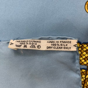 May include: A white tag with black text that reads "FABRIQUE EN FRANCE 100% SOIE" and "MADE IN FRANCE 100% SILK DRY CLEAN ONLY". The tag has a small circle with a letter "F" inside and a small triangle with a letter "A" inside. The tag is attached to a light blue fabric.