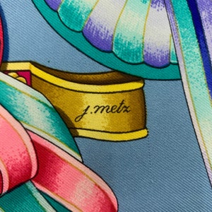 May include: A close-up of a blue and white silk scarf with a gold and yellow trim. The scarf features a floral design with a pink ribbon and a butterfly. The signature "J. Metz" is visible on the scarf.
