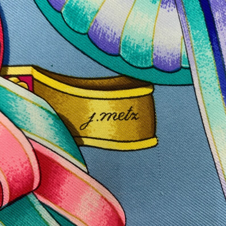 May include: A close-up of a blue and white silk scarf with a gold and yellow trim. The scarf features a floral design with a pink ribbon and a butterfly. The signature "J. Metz" is visible on the scarf.