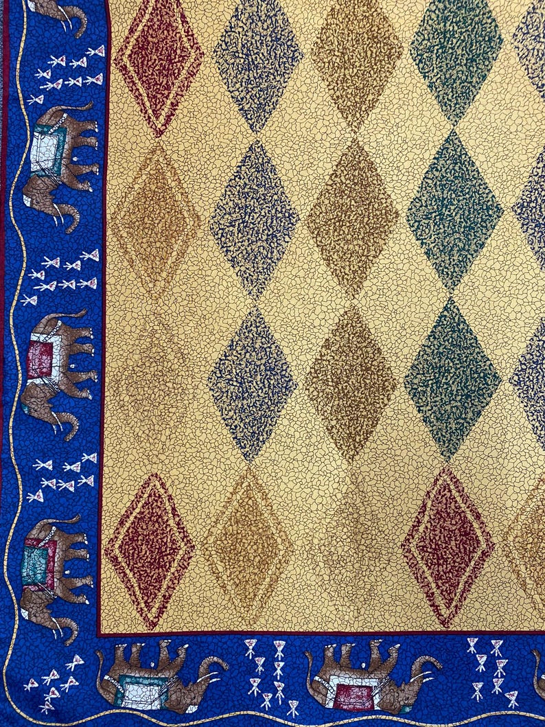 May include: A fabric textile featuring a cream-colored background with a repeating diamond pattern in shades of red, blue, and green. A blue border runs along the edge, decorated with a repeating motif of brown elephants and small white symbols.