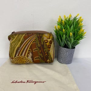 May include: A small, rectangular handbag with a brown leather top and a patterned fabric body. The fabric features a design with shades of brown, green, and yellow. A white cloth bag with the brand name "Salvatore Ferragamo" is in front.