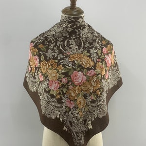 以下が含まれることがあります： ピンク、イエロー、ブラウンの花柄が施された、ブラウンのシルクスカーフ。白いペイズリー柄の縁取りが施されています。スカーフは白いマネキンに飾られています。スカーフの左下隅にブランド名「Lancel」が見えます。