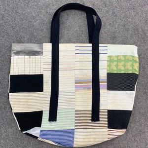 Op de afbeelding: Een patchwork tote bag met zwarte hengsels. De tas is gemaakt van verschillende stoffen met strepen, stippen en bloemenpatronen in tinten wit, zwart, bruin, blauw en groen.