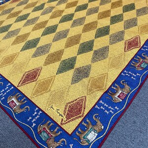 May include: A square scarf featuring a yellow field with a repeating diamond pattern in shades of green, brown, and red. The border is blue with a repeating elephant motif and small white symbols. The signature Jim Thompson is printed in black.