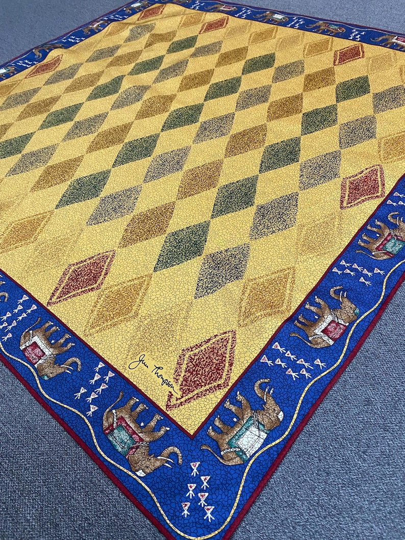 May include: A square scarf featuring a yellow field with a repeating diamond pattern in shades of green, brown, and red. The border is blue with a repeating elephant motif and small white symbols. The signature Jim Thompson is printed in black.