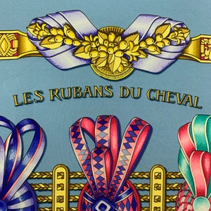 May include: A blue silk scarf with a gold and yellow design featuring a floral motif and the text "LES RUBANS DU CHEVAL". The scarf also features three colourful ribbon rosettes with geometric patterns.
