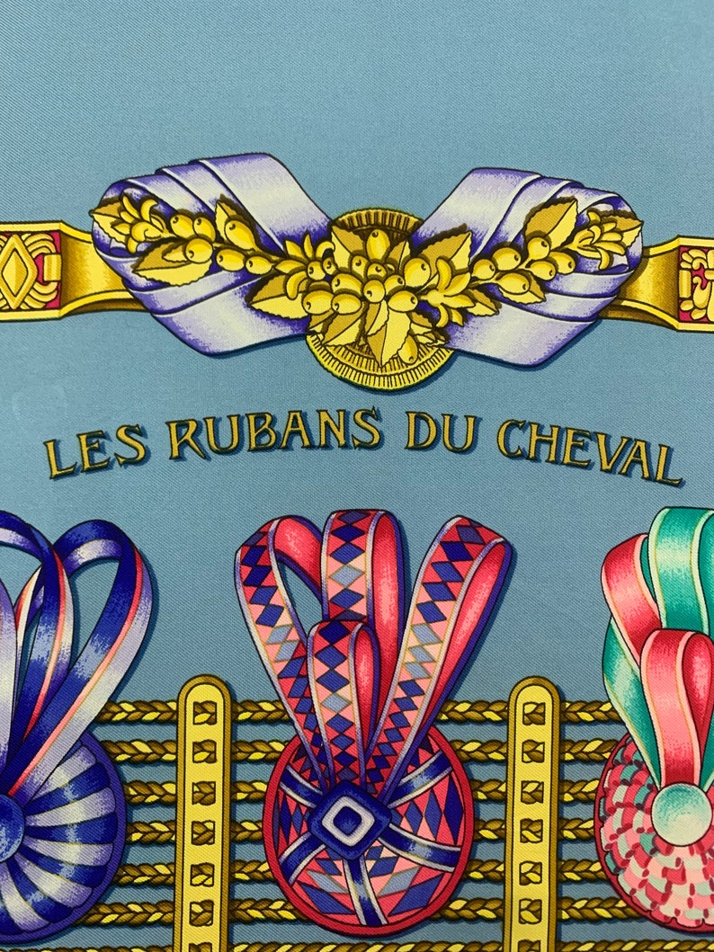 May include: A blue silk scarf with a gold and yellow design featuring a floral motif and the text "LES RUBANS DU CHEVAL". The scarf also features three colourful ribbon rosettes with geometric patterns.