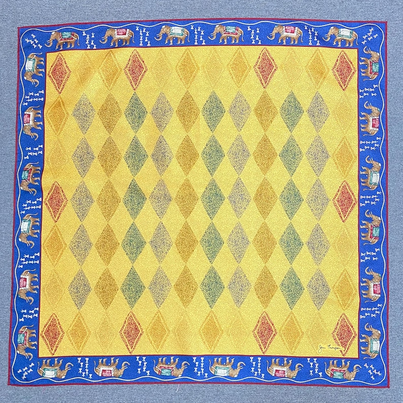 May include: A square scarf featuring a yellow field with a repeating diamond pattern in shades of gold, green, and red. The border is a deep blue with a repeating motif of elephants in profile, each wearing a decorative saddle, and small white icons.