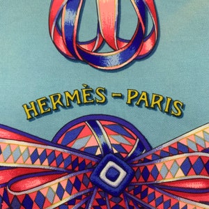 May include: A blue and pink patterned silk scarf with the text "HERM&Egrave;S - PARIS" in yellow. The scarf features a blue and pink ribbon design.