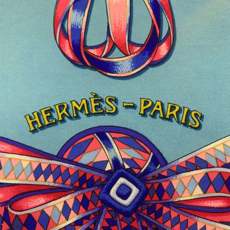 May include: A blue and pink patterned silk scarf with the text "HERM&Egrave;S - PARIS" in yellow. The scarf features a blue and pink ribbon design.