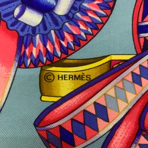 May include: A close-up of a blue and pink patterned fabric with a gold metal tag that reads "&copy; HERMES".