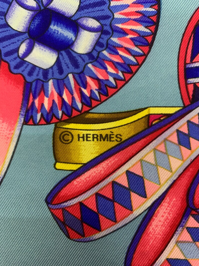 May include: A close-up of a blue and pink patterned fabric with a gold metal tag that reads "&copy; HERMES".
