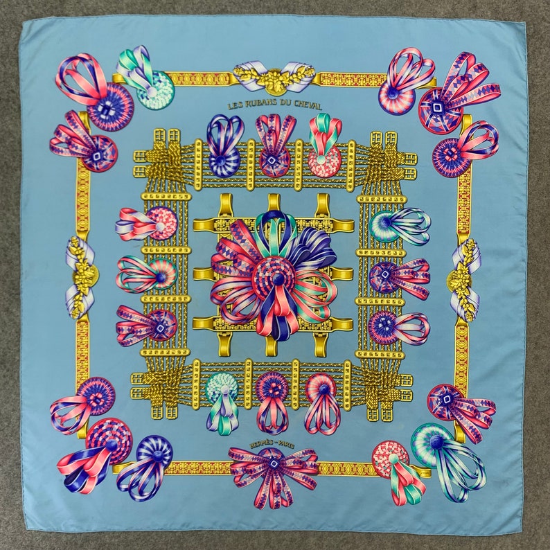 May include: A blue silk scarf with a gold and white pattern of ribbons and bows. The text "Les Rubans du Cheval" is printed in the centre of the scarf.