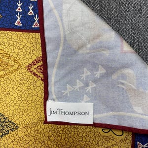 May include: A Jim Thompson scarf featuring a yellow section with a black crackle pattern, a blue section with white teepee-like motifs, and a light blue section with faint white illustrations. The scarf has a dark red border and a white brand label.