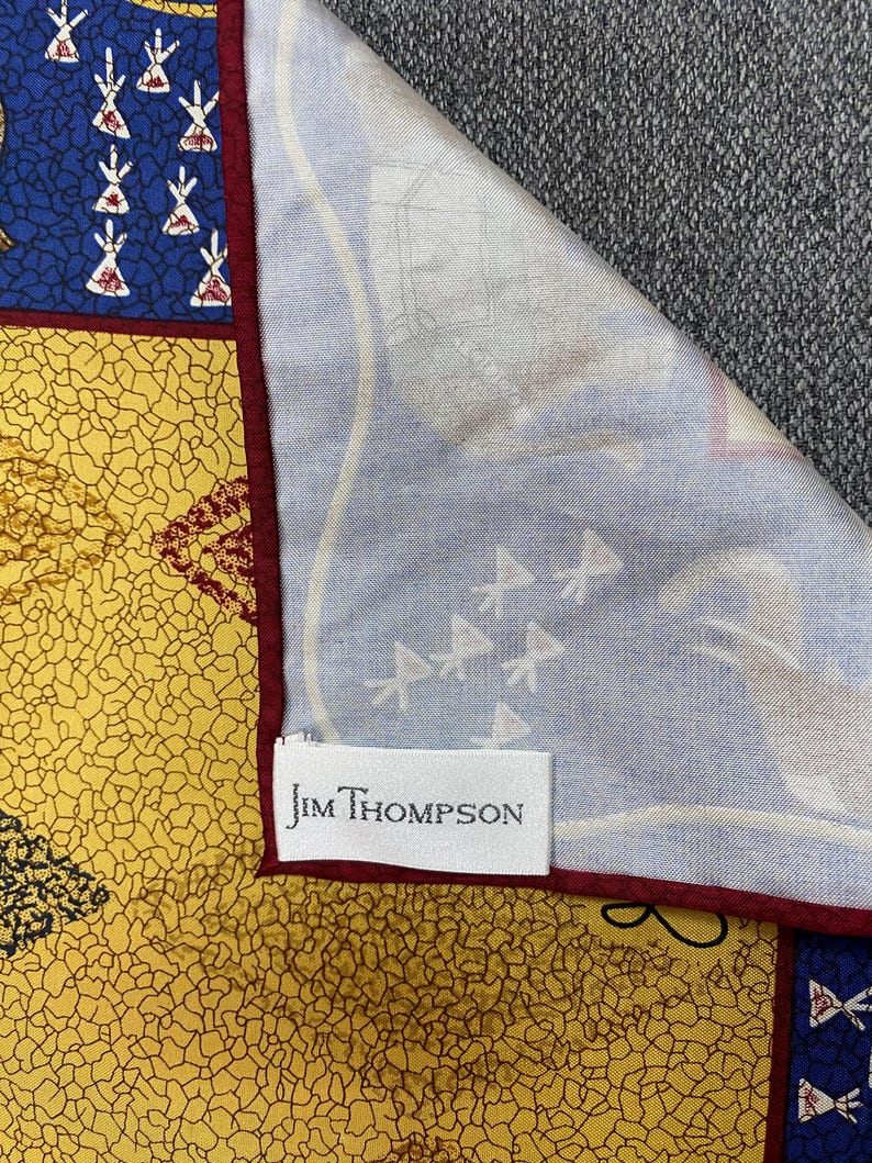 May include: A Jim Thompson scarf featuring a yellow section with a black crackle pattern, a blue section with white teepee-like motifs, and a light blue section with faint white illustrations. The scarf has a dark red border and a white brand label.
