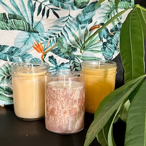 May include: Three candles in glass jars with different colored wax. The candles are on a dark surface with a tropical print fabric and a green plant in the background.