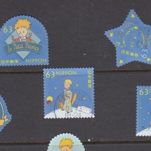 May include: A selection of vintage postage stamps depicting illustrations from "Le Petit Prince." The stamps are predominantly blue, showcasing images of the Little Prince, stars, and text in English and Japanese. Each stamp is valued at 63.