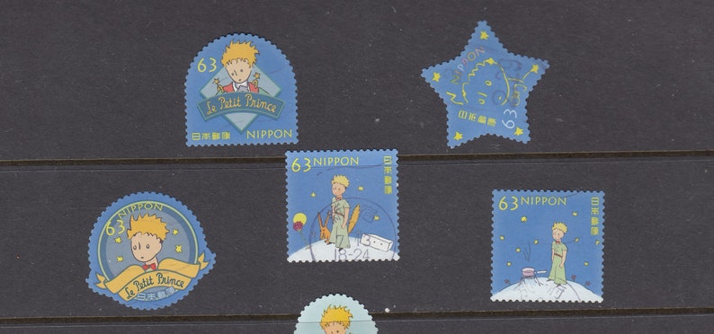 May include: A selection of vintage postage stamps depicting illustrations from "Le Petit Prince." The stamps are predominantly blue, showcasing images of the Little Prince, stars, and text in English and Japanese. Each stamp is valued at 63.