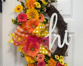 May include: A wreath with a brown vine base decorated with yellow, orange, and pink flowers. A white wooden sign with the word "hi" is attached to the wreath. A yellow and white polka dot ribbon with orange and pink checkered ribbon is tied around the base of the wreath.