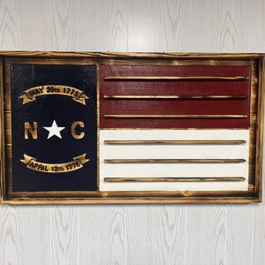 May include: A wooden North Carolina flag with a red, white, and blue color scheme. The flag features the state seal with the text "May 20th 1775" and "April 12th 1776" and the letters "N.C." with a star in the center. The flag is mounted in a wooden frame.