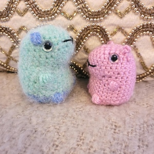May include: Two small, handmade crochet creatures. One is light green with blue accents, the other is pink. Both have black eyes and small tails. They are sitting on a textured, light-colored surface, with a beaded background.