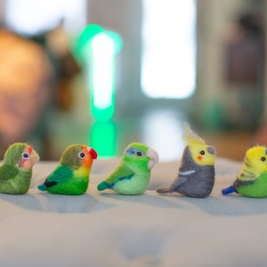 May include: Five felt bird ornaments in a row, featuring green, yellow, and grey plumage. The birds have detailed beaks and wings. They are displayed on a soft, light-coloured surface.