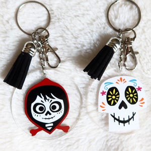 Puede incluir: Dos llaveros con borlas negras. Un llavero presenta un dije redondo de acrílico transparente con una imagen de dibujos animados de Miguel de la película Coco. El otro llavero presenta un dije redondo de acrílico transparente con un diseño de calavera de azúcar blanca.