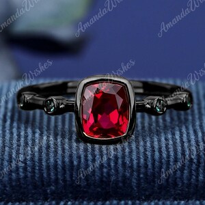 Può includere: Un anello nero con una pietra rossa quadrata e due pietre verdi più piccole su ogni lato. L'anello è su una scatola di velluto blu.
