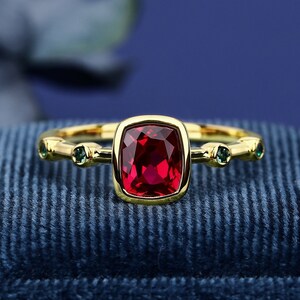 May include: A gold ring featuring a red gemstone and green accent stones. The ring is displayed on a blue velvet box.