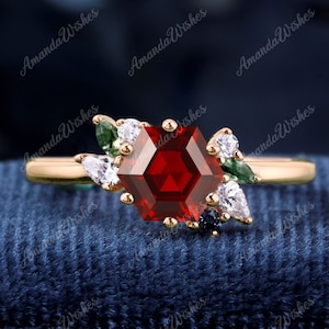 May include: A gold ring featuring a red hexagonal gemstone surrounded by smaller white and green gemstones. The ring is displayed on a blue velvet background.
