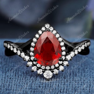 Può includere: Un anello nero con una pietra rossa a forma di pera circondata da piccole pietre bianche. L'anello ha un design delicato e intricato con una montatura a halo.