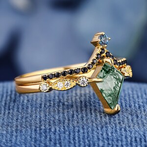 Op de afbeelding: Een gouden ring met een groene, blauwe en zwarte edelsteen. De ring ligt op een blauwe stoffen achtergrond.