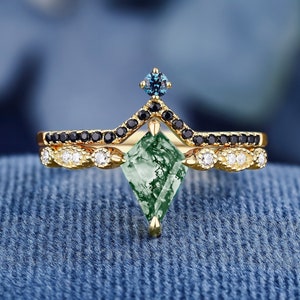 Puede incluir: Un anillo de oro con una piedra preciosa de ágata verde musgo, diamantes negros y un acento de zafiro azul. El anillo está colocado sobre un fondo de mezclilla azul.