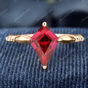 May include: A rose gold ring with a square-shaped, red gemstone set in the center. The ring has a delicate design with curved accents on either side of the stone.