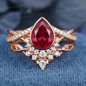 Può includere: Un anello in oro rosa con una gemma rossa centrale a forma di pera circondata da un gruppo di piccole pietre bianche rotonde. La fascia presenta un design diviso e intrecciato con ulteriori piccole pietre bianche incastonate sulla parte superiore.