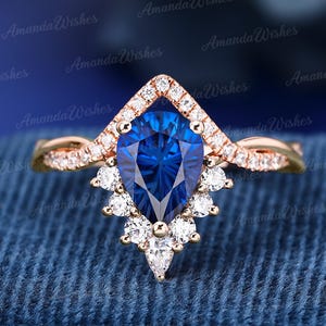 Può includere: Un anello in oro rosa con una pietra preziosa blu scuro a forma di pera come punto focale. L'anello presenta un'aureola a forma di V di piccole pietre chiare e una fascia intrecciata. Ulteriori piccole pietre chiare sono incastonate sotto.