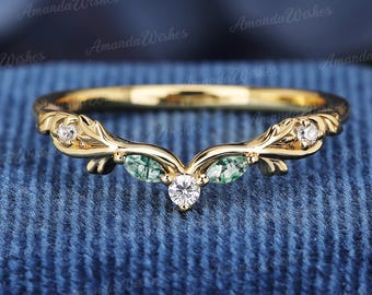 May include: A gold-colored ring with a curved design. The ring features small, clear gemstones and green stones. The ring has a leaf-like pattern on the sides and is set against a blue background.
