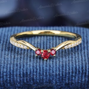 May include: A gold chevron-shaped ring featuring three central round-cut red rubies and a series of small clear white accent stones set along the band. The metal band has a polished finish and a distinct V-shaped curve at the centre point.