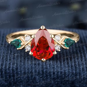 Peut inclure: Une bague en or avec une pierre rouge en forme de poire au centre, encadrée de deux pierres vertes émeraude. De petites pierres blanches ajoutent de l'éclat. L'ensemble est posé sur un fond bleu foncé.